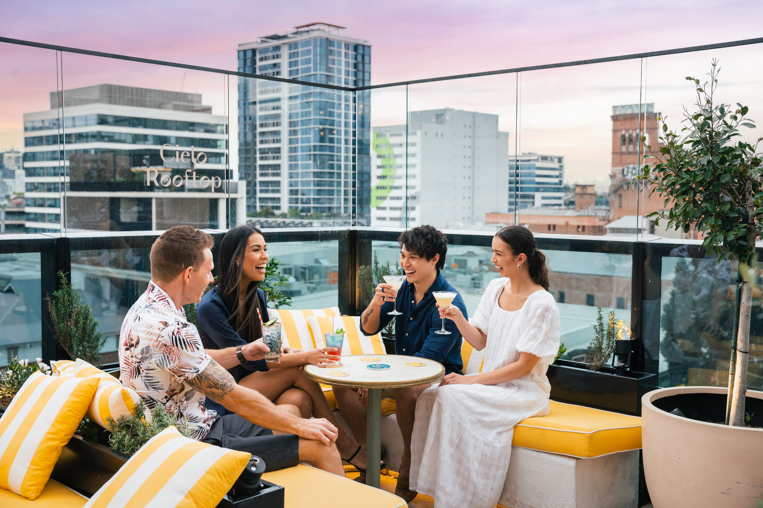 Four friends relax on yellow sun lounges sipping vibrant cocktails at Cielo Rooftop Bar
