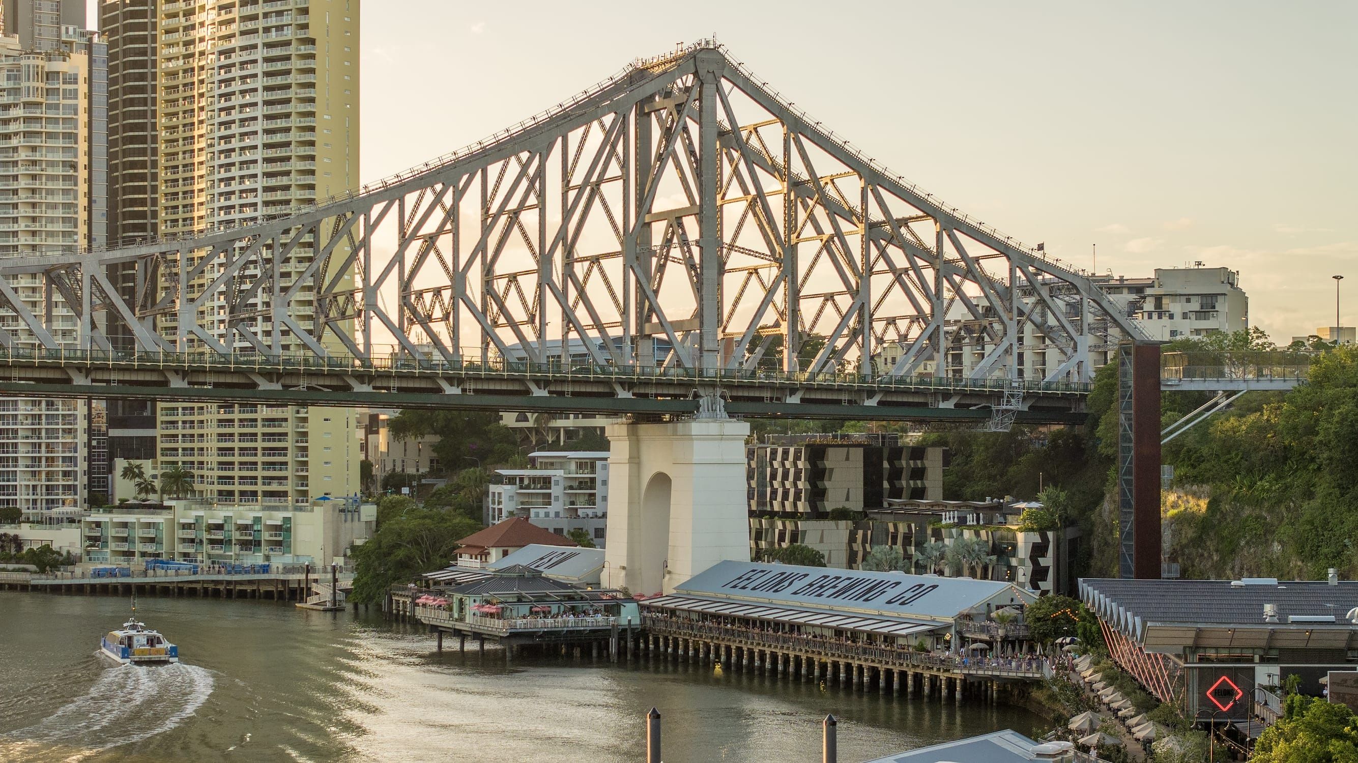 Aerial view of riverfront precinct Howard Smith Wharves, Story Bridge and city skyline as a CityCat ferry travels up the river