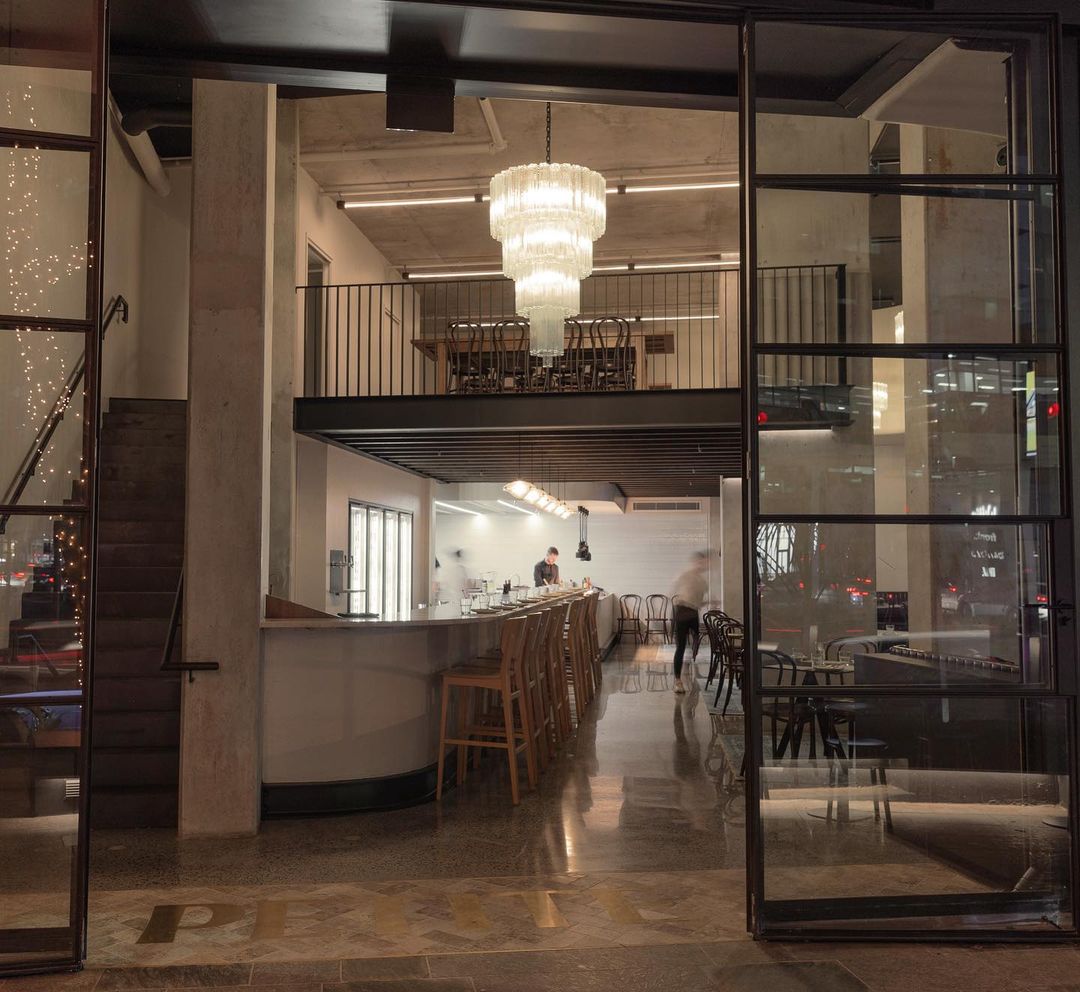 View of Petite bistro and wine bar through tall glass entranceway, with vintage chandelier hanging from ceiling View of Petite bistro and wine bar through tall glass entranceway, with vintage chandelier hanging from ceiling