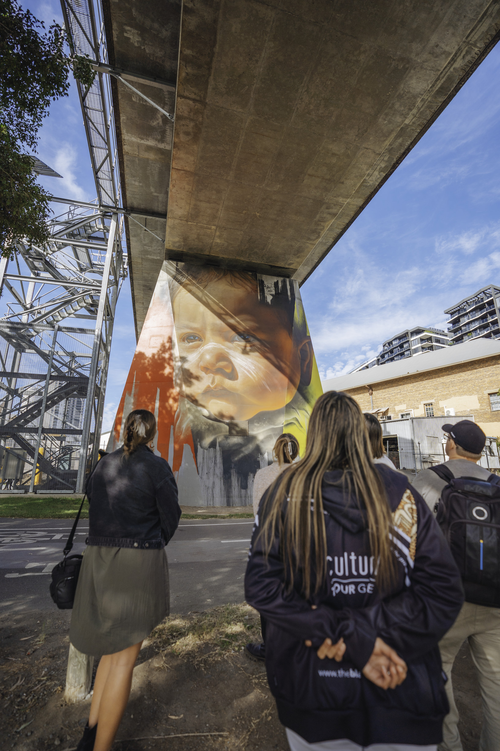 Group experiencing Indigenous walking tour in Brisbane with Blackcard Cultural Tours. Pictured, the group is standing under a city bridge to discover a hidden street artwork depicting a young Aboriginal child Group experiencing Indigenous walking tour in Brisbane with Blackcard Cultural Tours. Pictured, the group is standing under a city bridge to discover a hidden street artwork depicting a young Aboriginal child