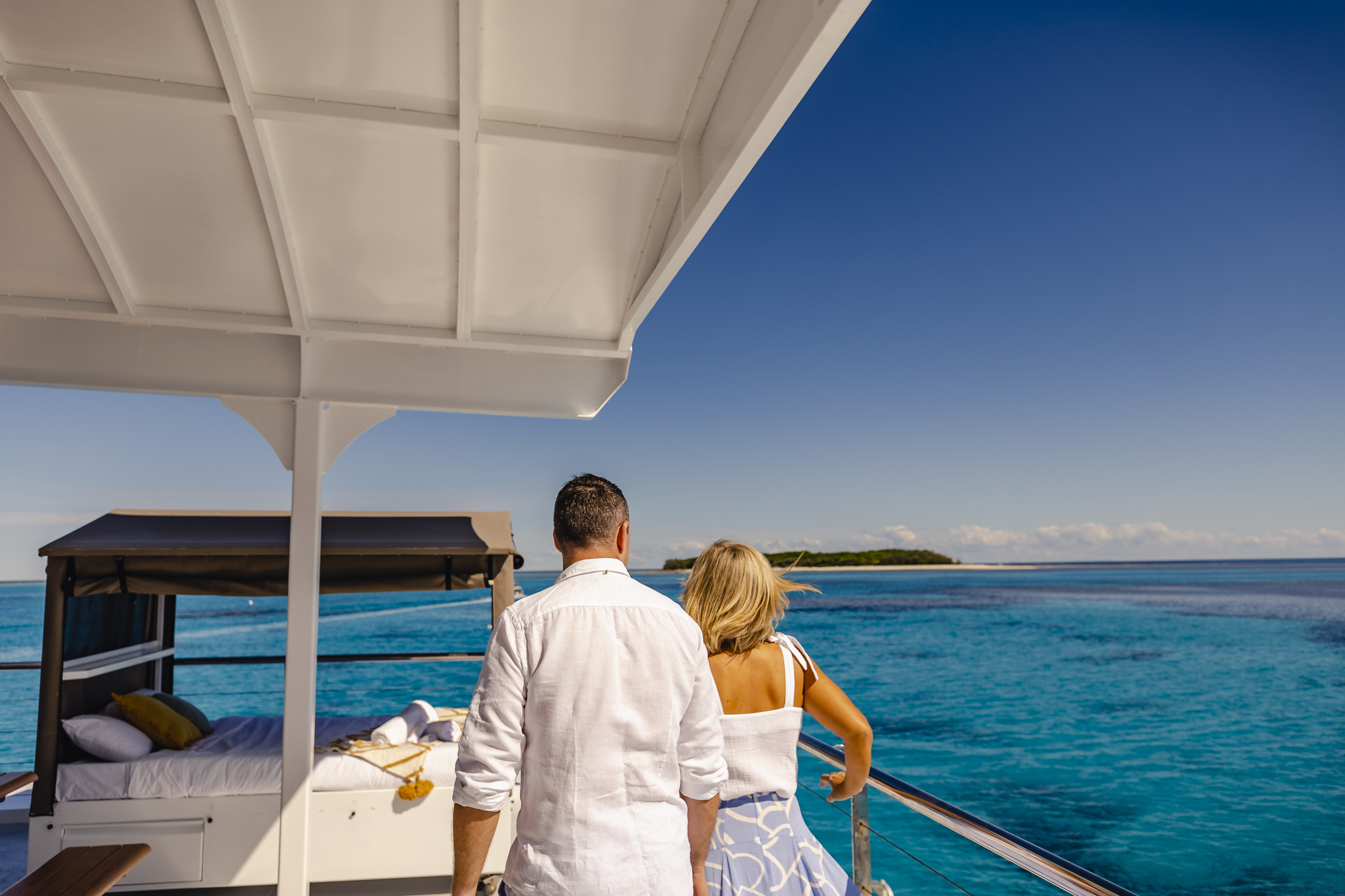Couple on the top deck of Lady Musgrave Experience HQ, with glamping experience and Lady Musgrave Island in the background