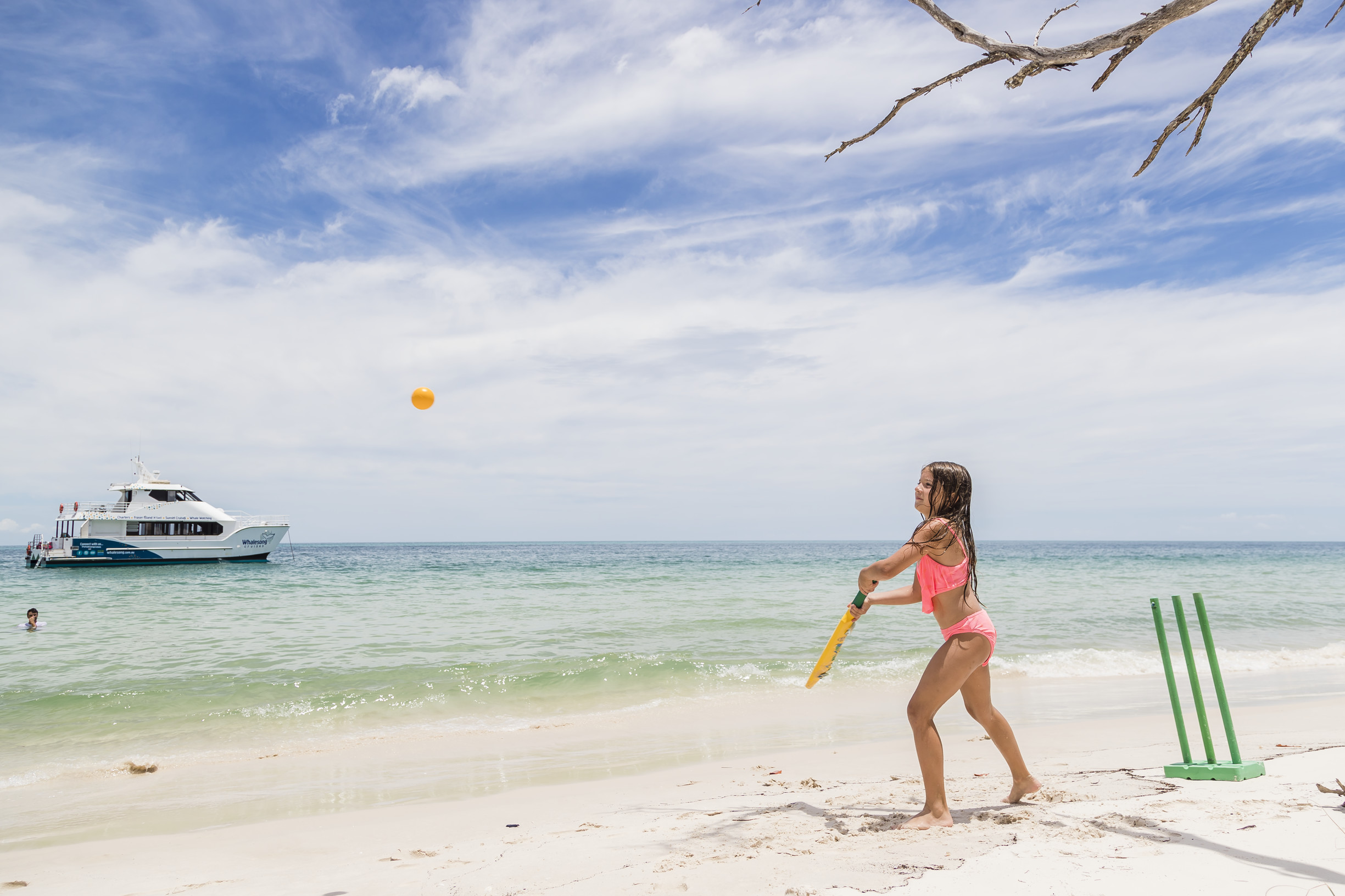 Child playing beach cricket on K'gari, with the Whalesong Cruises boat in the background