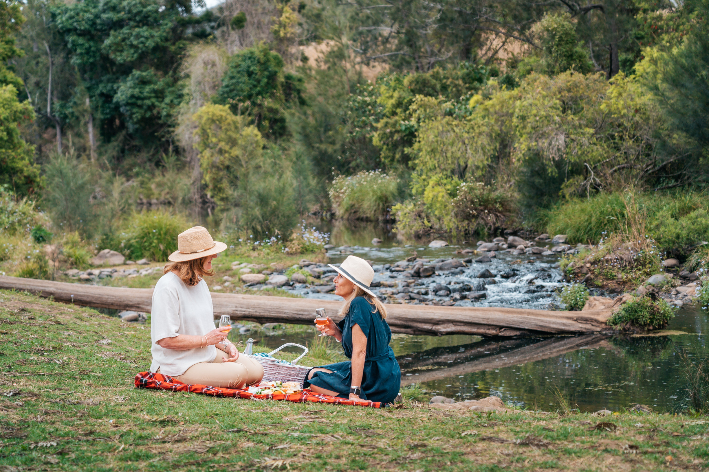 Two ladies sitting on picnic rug by the river sipping wine