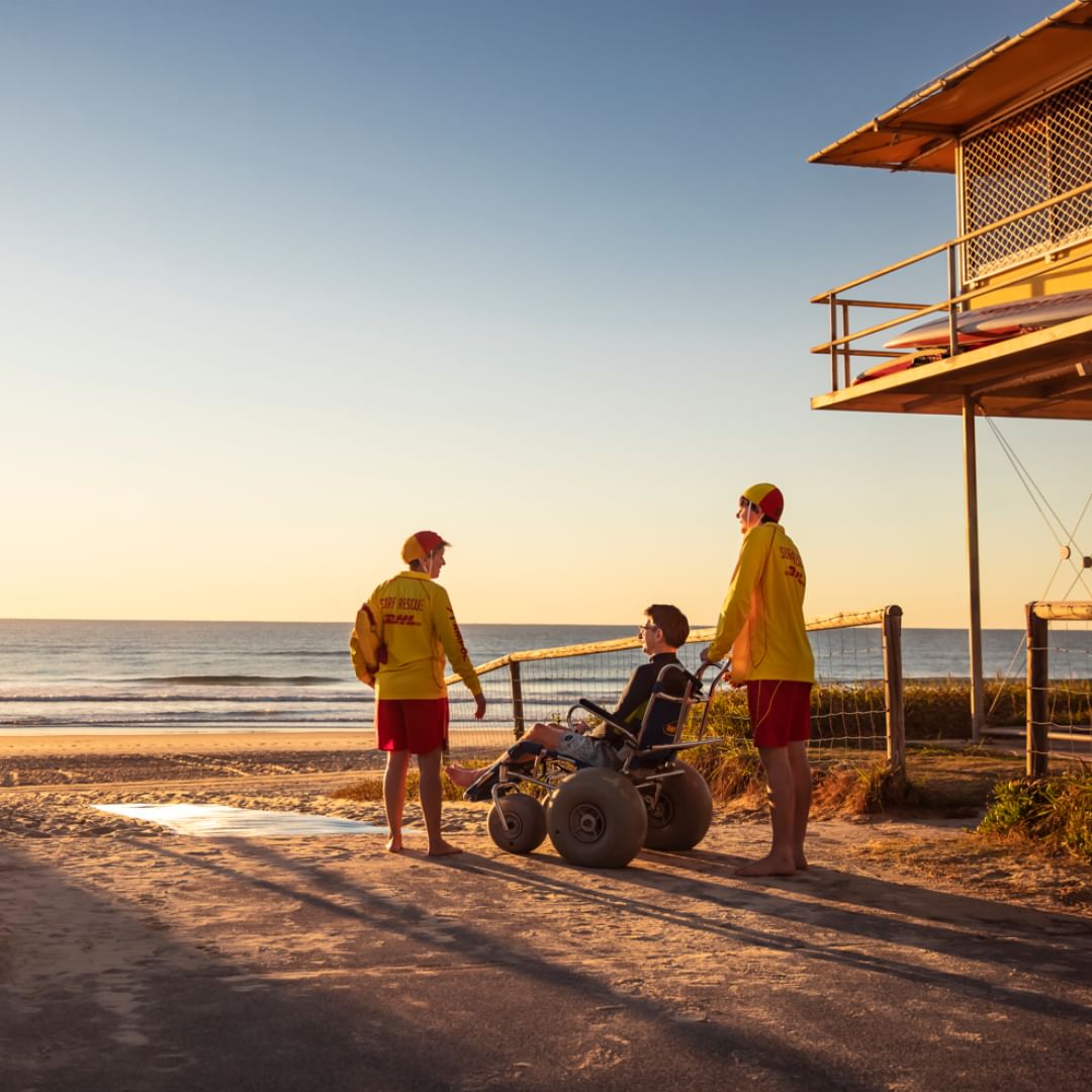 Person in a beach wheelchair at a Gold Coast beach, accompanied by two lifeguards Person in a beach wheelchair at a Gold Coast beach, accompanied by two lifeguards
