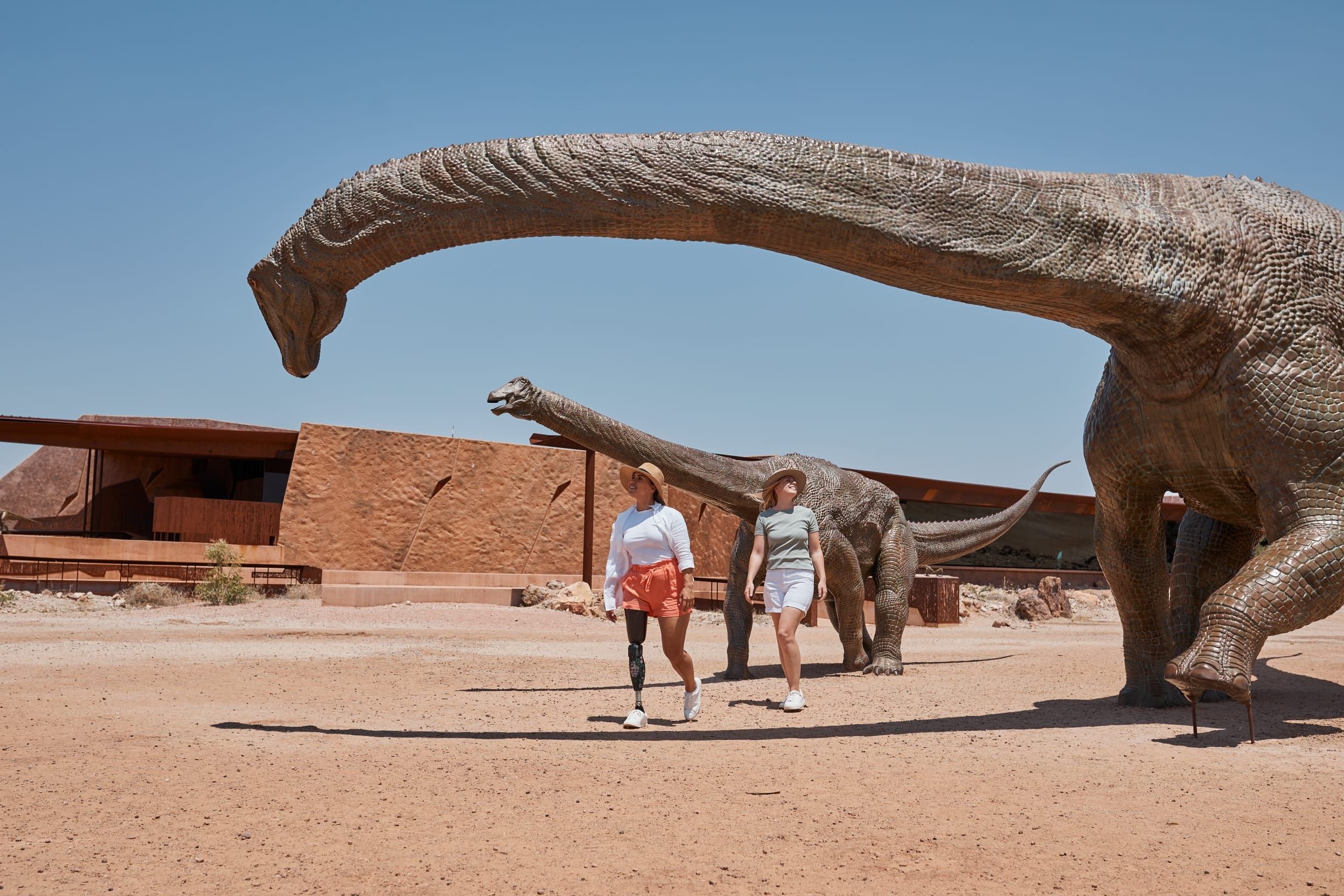 Two women walk under replica dinosaurs outside Australian Age of Dinosaurs museum in Winton