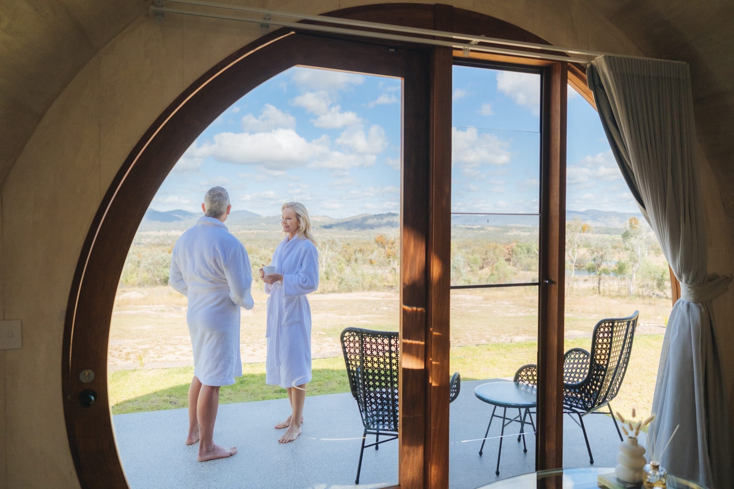 Couple in bathrobes stand on the deck of their wine-barrel shaped cabin in the countryside