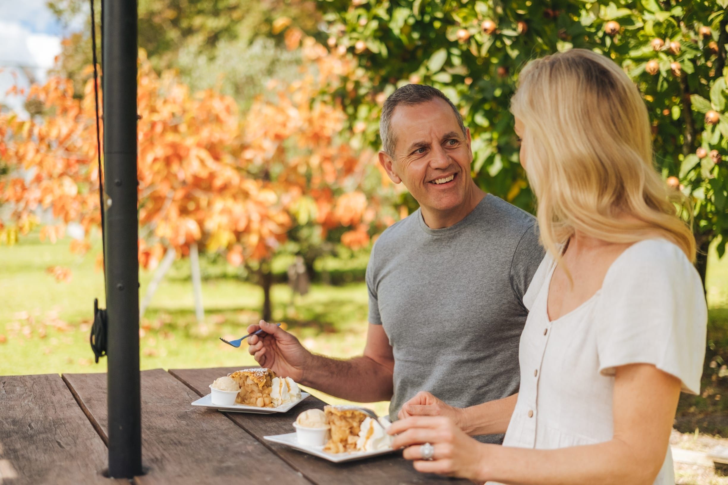 A couple sitting down, eating apple pie, with orange leaves in the background