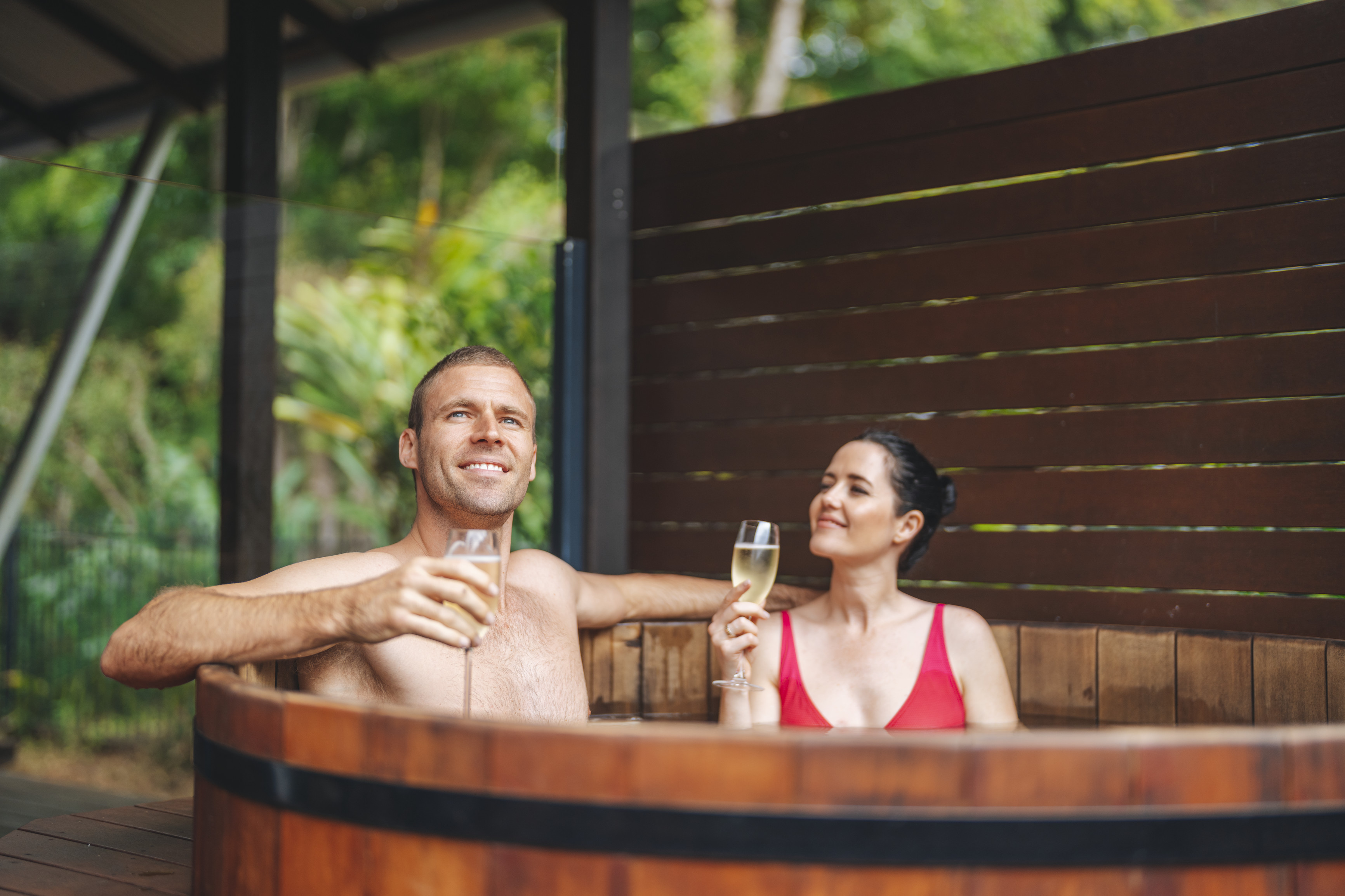 Couple relaxing in a cedar hot tub on the deck of their accommodation, champagne in hand