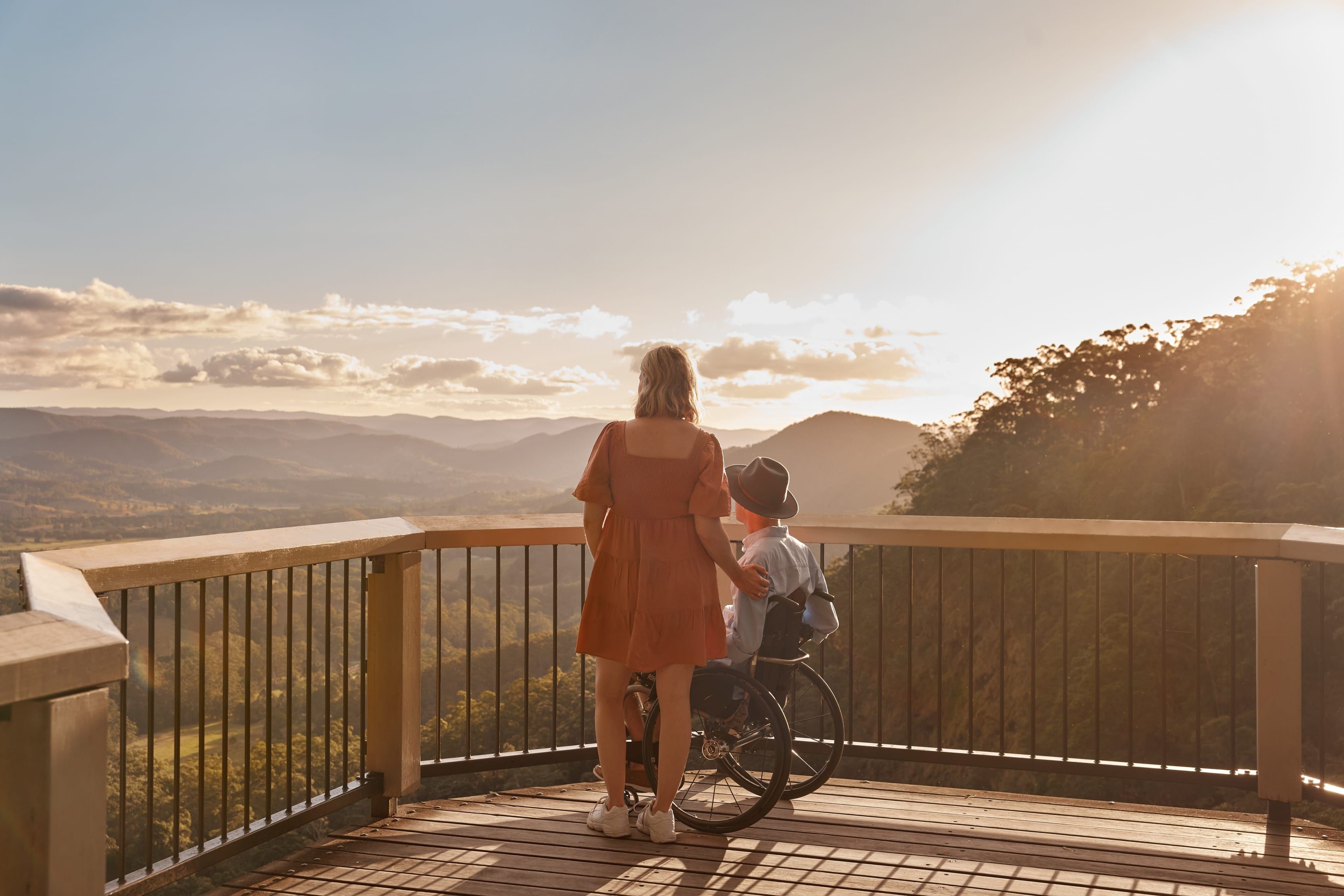Man in wheelchair and woman watch sunset over a mountain range from an accessible lookout