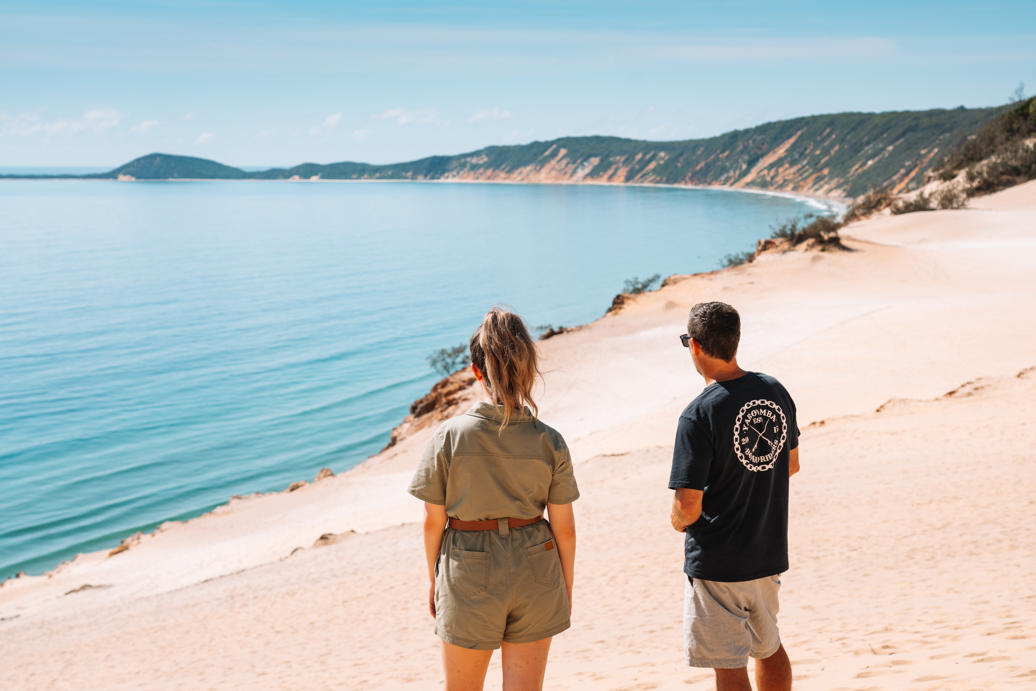 Man and woman stand at the top of a giant sand dune overlooking the beach and ocean