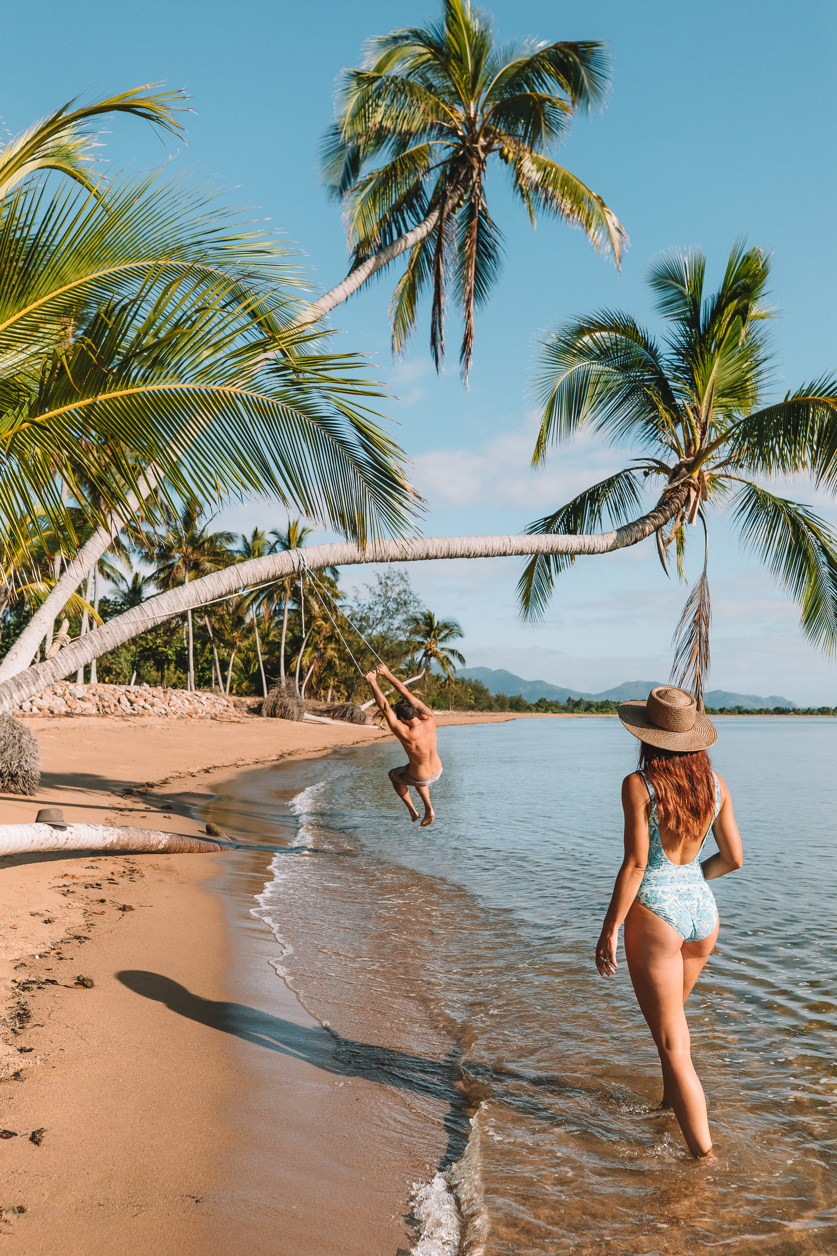Man swinging on a palm tree over Lucinda Beach, while woman walks on the sand nearby