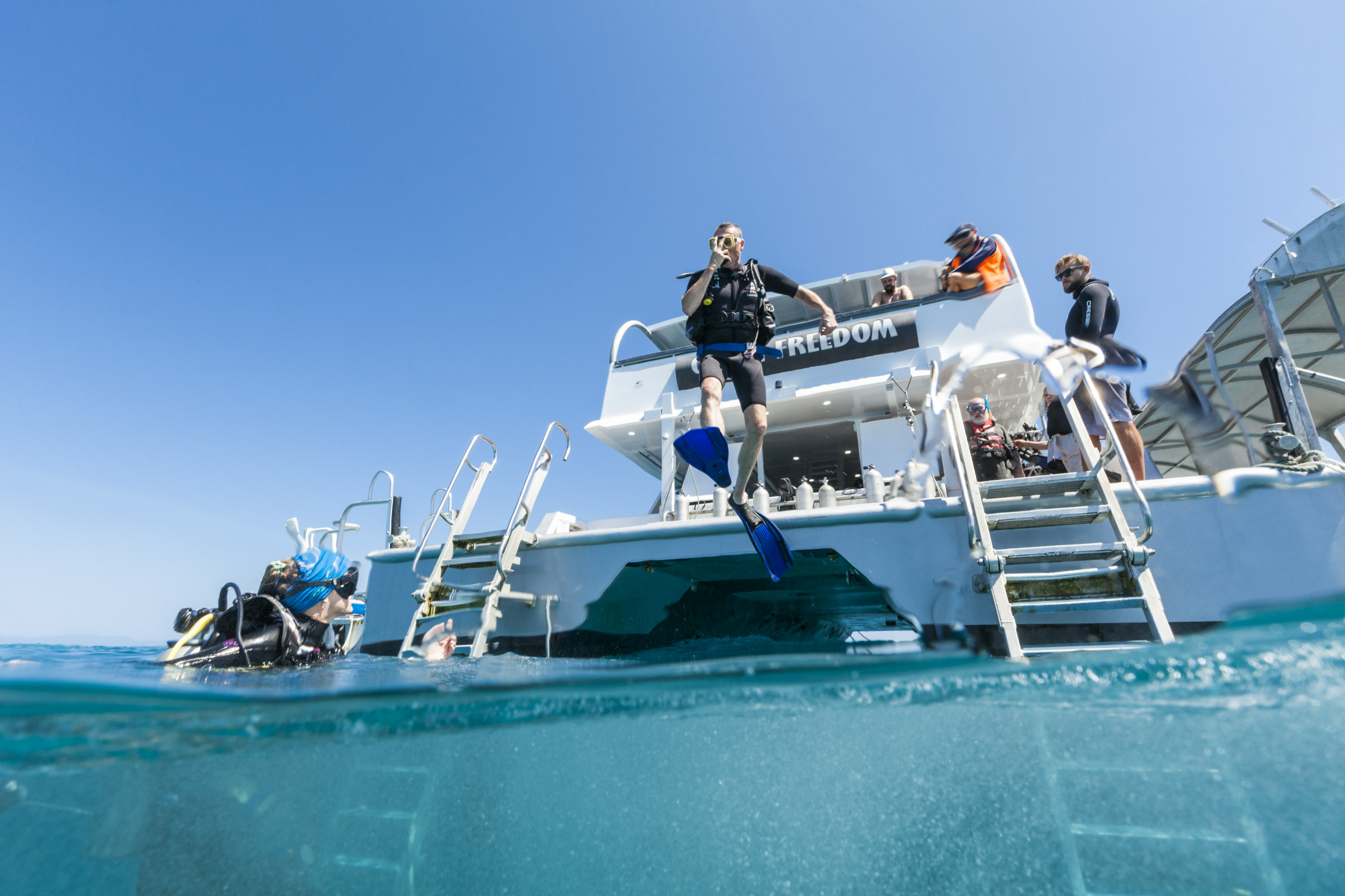An introductory diver leaps into the water from the back deck of the Ocean Freedom boat, with the dive instructor watching, Ocean Freedom | scuba diving great barrier reef first time An introductory diver leaps into the water from the back deck of the Ocean Freedom boat, with the dive instructor watching, Ocean Freedom | scuba diving great barrier reef first time