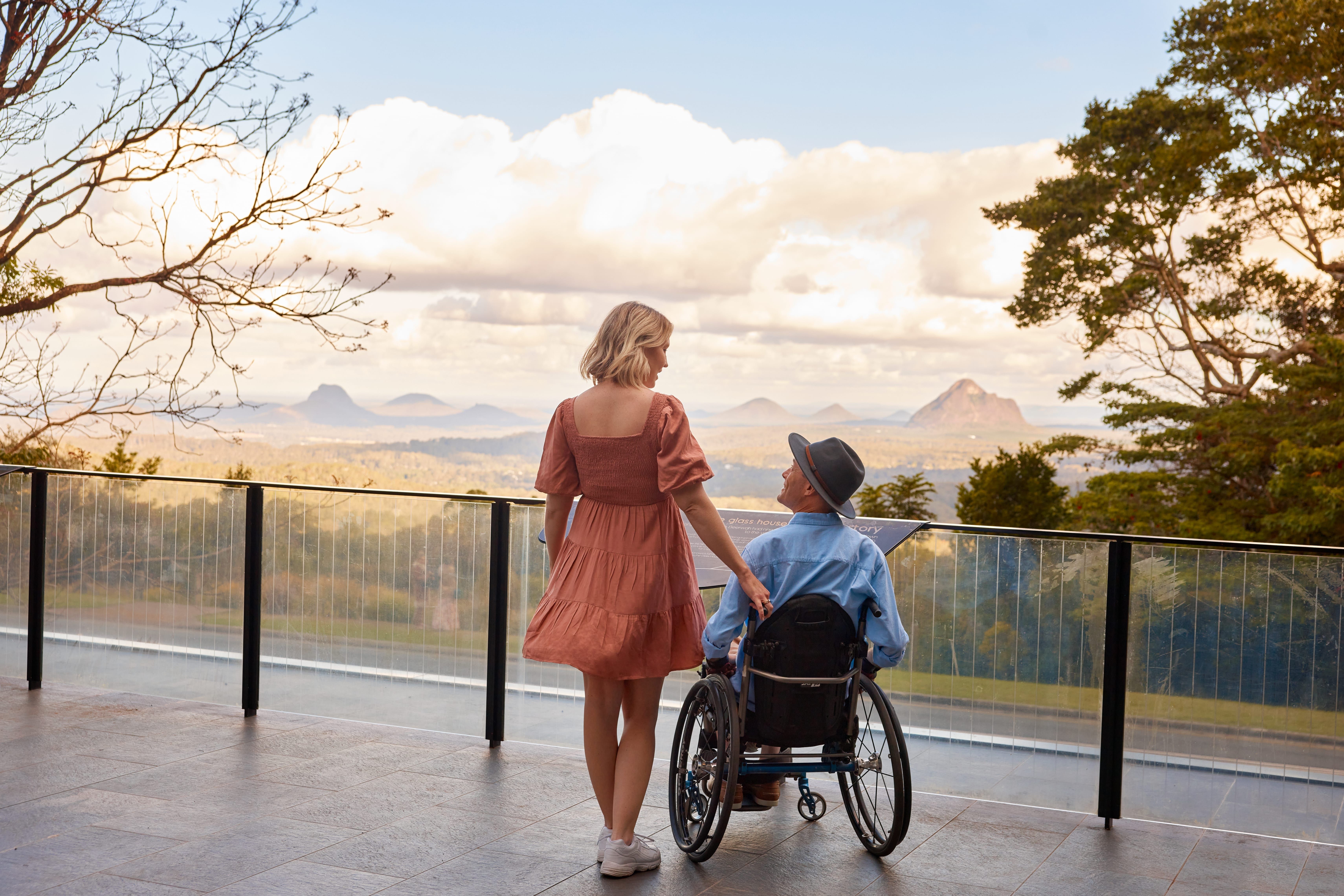 Man in wheelchair and woman at a scenic lookout that looks over the peaks of the Glass House Mountains Man in wheelchair and woman at a scenic lookout that looks over the peaks of the Glass House Mountains