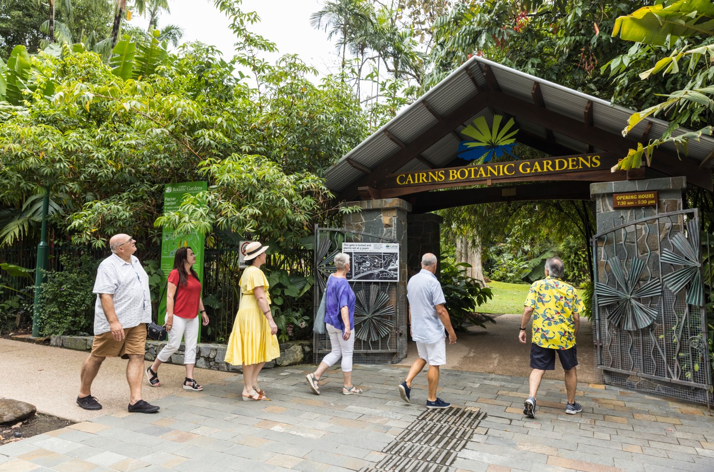 A group of travellers are apporaching the Cairns Botanic Gardens entry that is surrounded by lush green plants and tall palm trees.